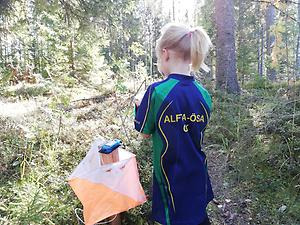 Ett barn i träningskläder står i skogen intill en orienteringsskärm.