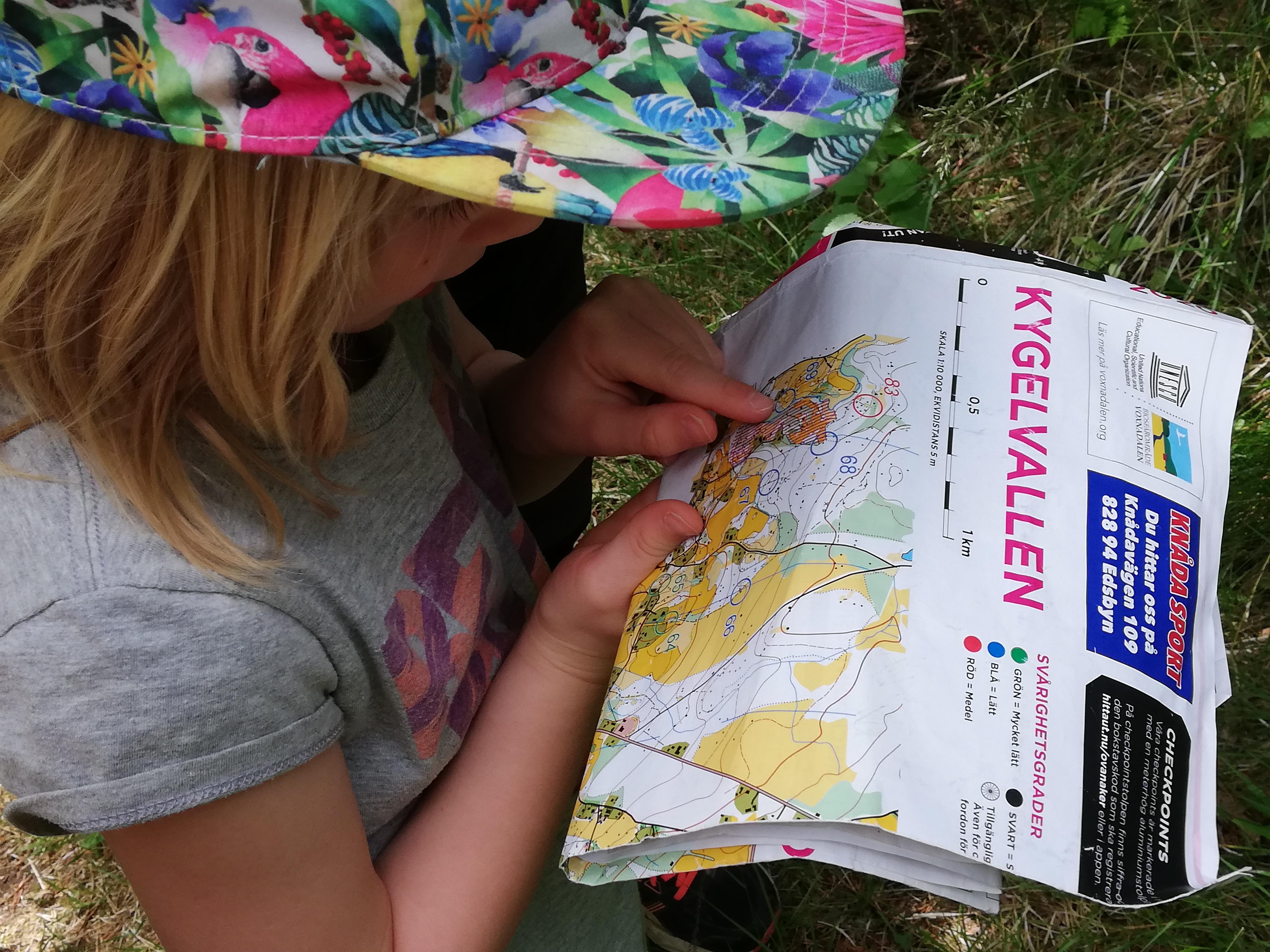 Ett barn i keps står ute i naturen och tittar ner på en orienteringskarta med rubriken "Kygelvallen".